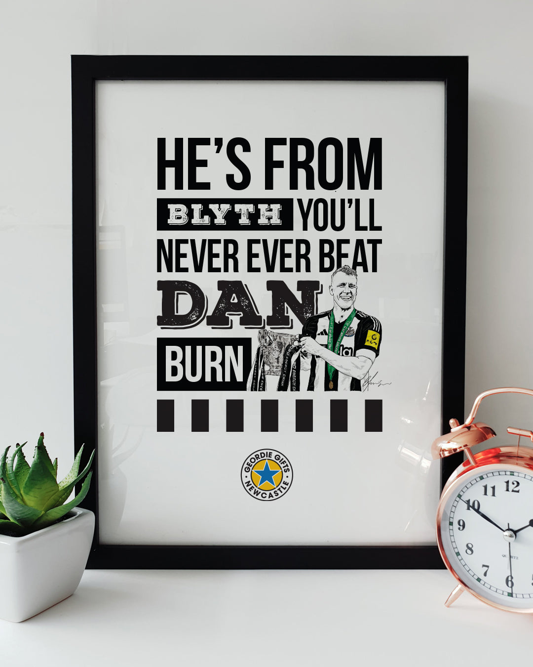 Geordie Gifts Newcastle United print for supporters, featuring a hand-drawn Dan Burn illustration with the Carabao Cup trophy and the fan chant ‘He’s from Blyth, you’ll never ever beat Dan Burn,’ displayed in a black frame with a plant and clock.