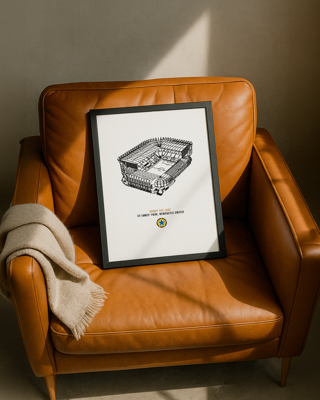A framed Geordie Gifts print of St. James' Park stadium leaning against a tan leather armchair.