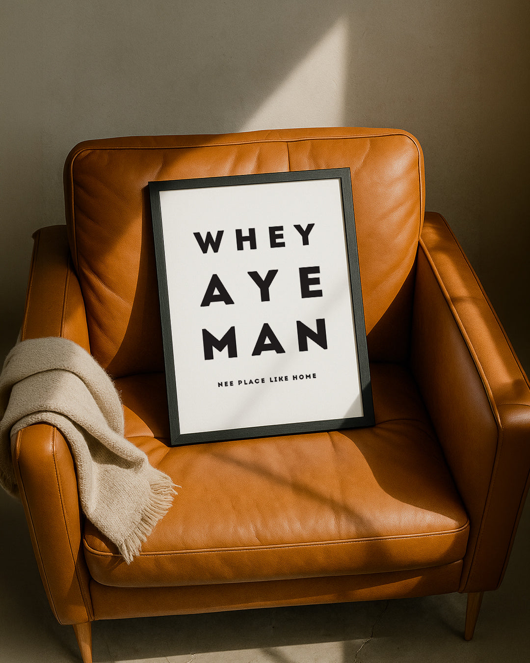 A black-framed "Whey Aye Man" typography poster leaning against a stylish tan leather armchair design by geordie gifts