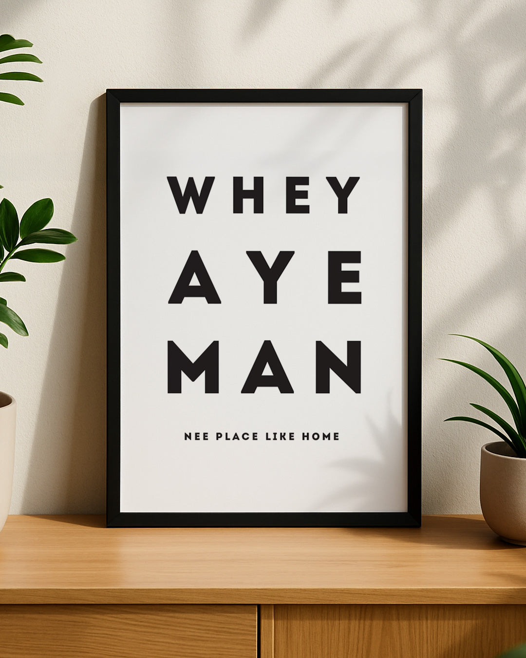 A framed Geordie Gifts print reading "Whey Aye Man - Nee Place Like Home" hanging on a neutral wall next to a green plant.