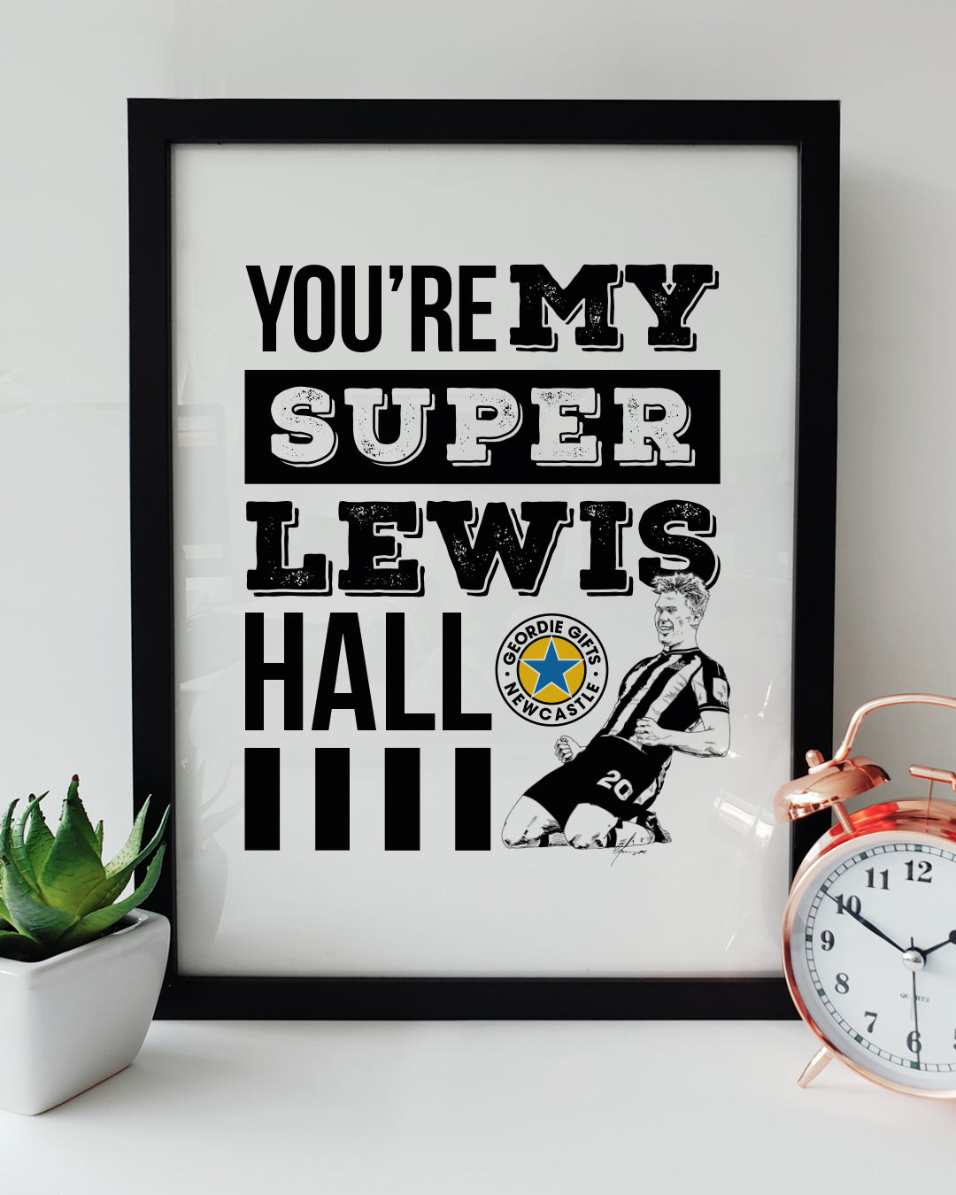Black framed "You're My Super Lewis Hall" Newcastle United chant print featuring Lewis Hall goal celebration illustration by Geordie Gifts next to a rose gold alarm clock.