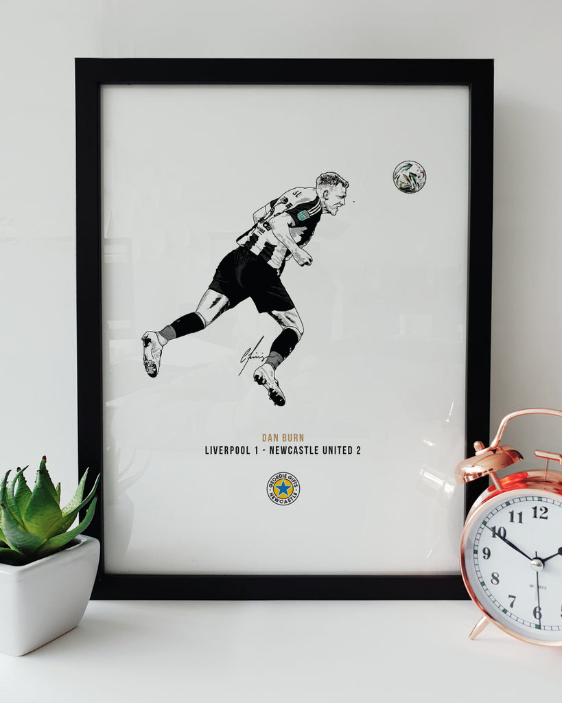 Geordie Gifts Newcastle United print for supporters, featuring a hand-drawn Dan Burn illustration scoring the first goal in the Carabao Cup final against Liverpool at Wembley, displayed in a black frame with a plant and clock.