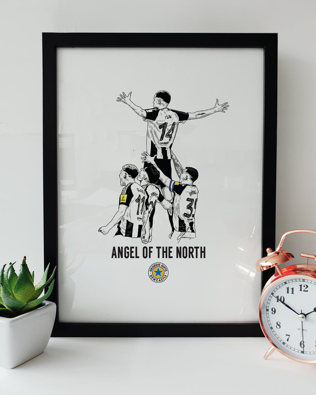 Geordie Gifts Newcastle United print featuring a hand-drawn Alexander Isak illustration as the Angel of the North, celebrating his Carabao Cup final goal against Liverpool, displayed in a black frame with a plant and clock.