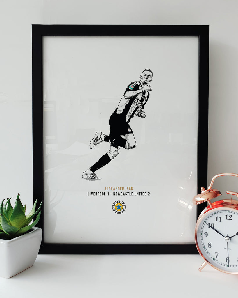 Geordie Gifts Newcastle United print for supporters, featuring a hand-drawn Alexander Isak illustration celebrating his Carabao Cup final goal against Liverpool by pointing at Virgil van Dijk, displayed in a black frame with a plant and clock.