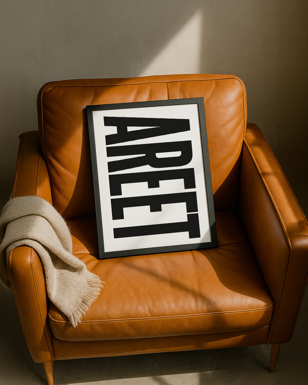 Framed "AREET" typography print resting casually on a tan leather armchair next to a folded beige throw blanket, illuminated by warm sunlight. Designed for Geordie Gifts.