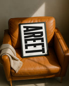 Framed "AREET" typography print resting casually on a tan leather armchair next to a folded beige throw blanket, illuminated by warm sunlight. Designed for Geordie Gifts.