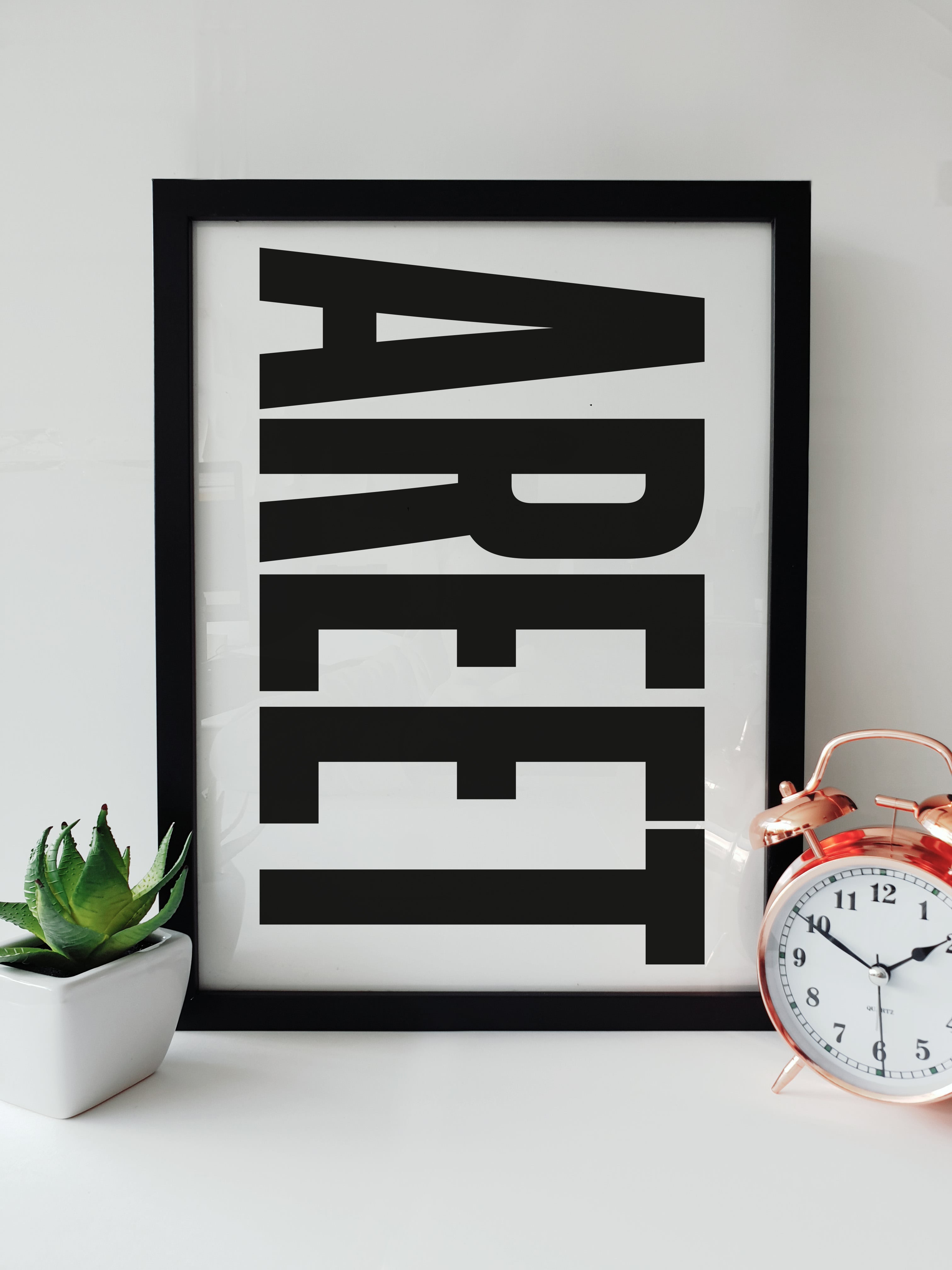 Framed "AREET" black and white typography print displayed on a clean white shelf next to a small potted succulent and a classic copper alarm clock. Designed for Geordie Gifts.