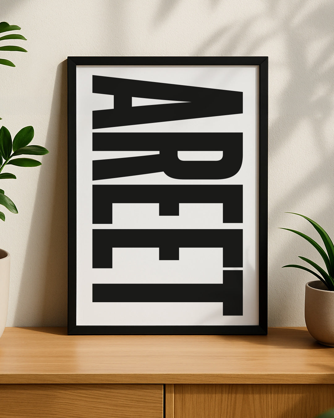 Framed bold "AREET" typography print resting on a modern wooden sideboard, styled with green house plants on either side with natural shadows cast on the wall behind it. Designed for Geordie Gifts.