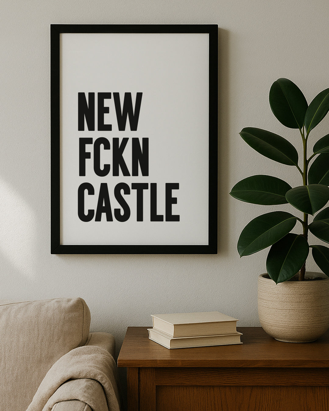 Framed vertical typography print featuring the words "NEW FCKN CASTLE" stacked in bold black capital letters on a white background. Displayed on a wooden table next to a large green rubber plant. Designed for Geordie Gifts.