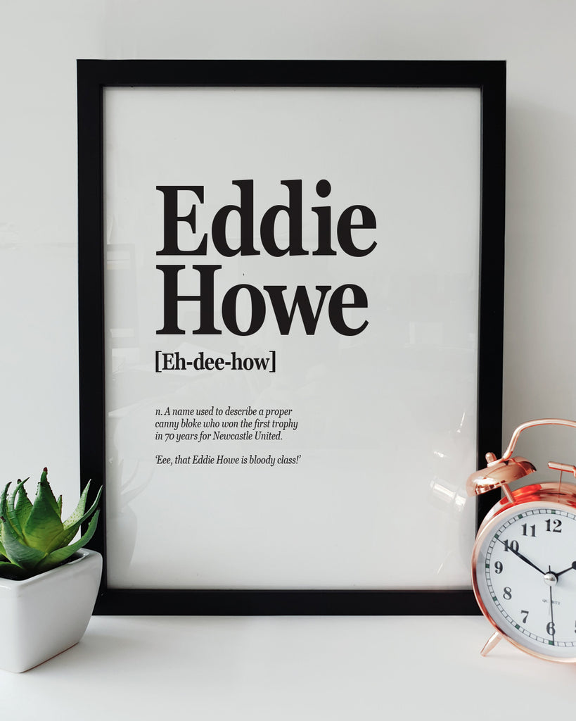 Geordie Gifts Newcastle United print for supporters, featuring a typographic design with the definition of Eddie Howe as the manager who won the first trophy in 70 years, displayed in a black frame with a plant and clock.