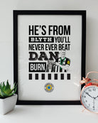 Geordie Gifts Newcastle United print for supporters, featuring a hand-drawn Dan Burn illustration with the Carabao Cup trophy and the fan chant ‘He’s from Blyth, you’ll never ever beat Dan Burn,’ displayed in a black frame with a plant and clock.