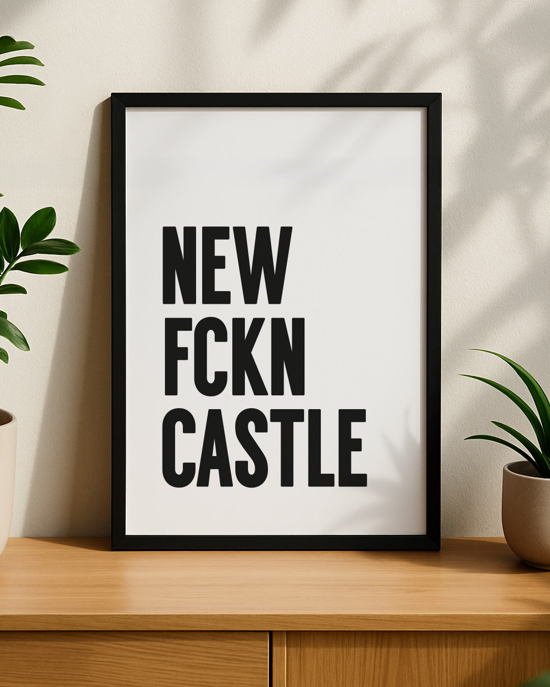 Framed bold "NEW FCKN CASTLE" typography print resting on a modern wooden sideboard, styled with green house plants on either side with natural shadows cast on the wall behind it. Designed for Geordie Gifts.
