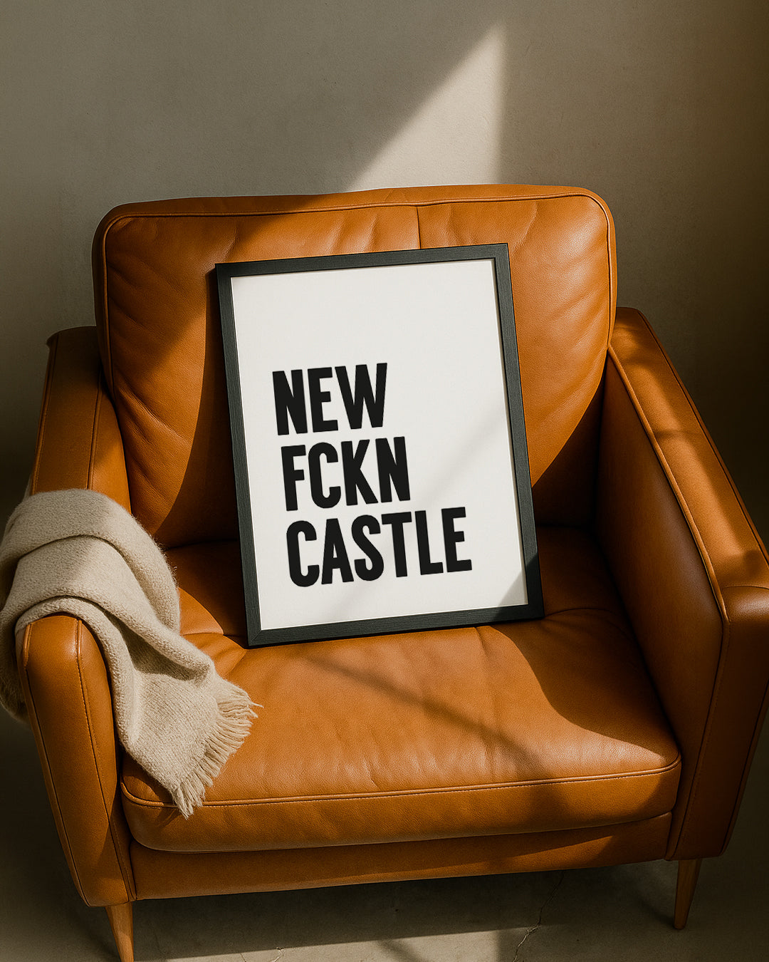 Framed "NEW FCKN CASTLE" typography print resting casually on a tan leather armchair next to a folded beige throw blanket, illuminated by warm sunlight. Designed for Geordie Gifts.