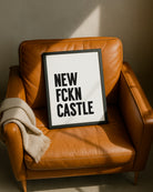 Framed "NEW FCKN CASTLE" typography print resting casually on a tan leather armchair next to a folded beige throw blanket, illuminated by warm sunlight. Designed for Geordie Gifts.
