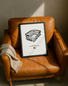 A framed Geordie Gifts print of St. James' Park stadium leaning against a tan leather armchair.