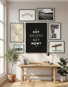 A black 'Shy Bairns Get Nowt' framed print on a wooden desk with a lamp and stationery.