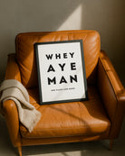 A black-framed "Whey Aye Man" typography poster leaning against a stylish tan leather armchair design by geordie gifts
