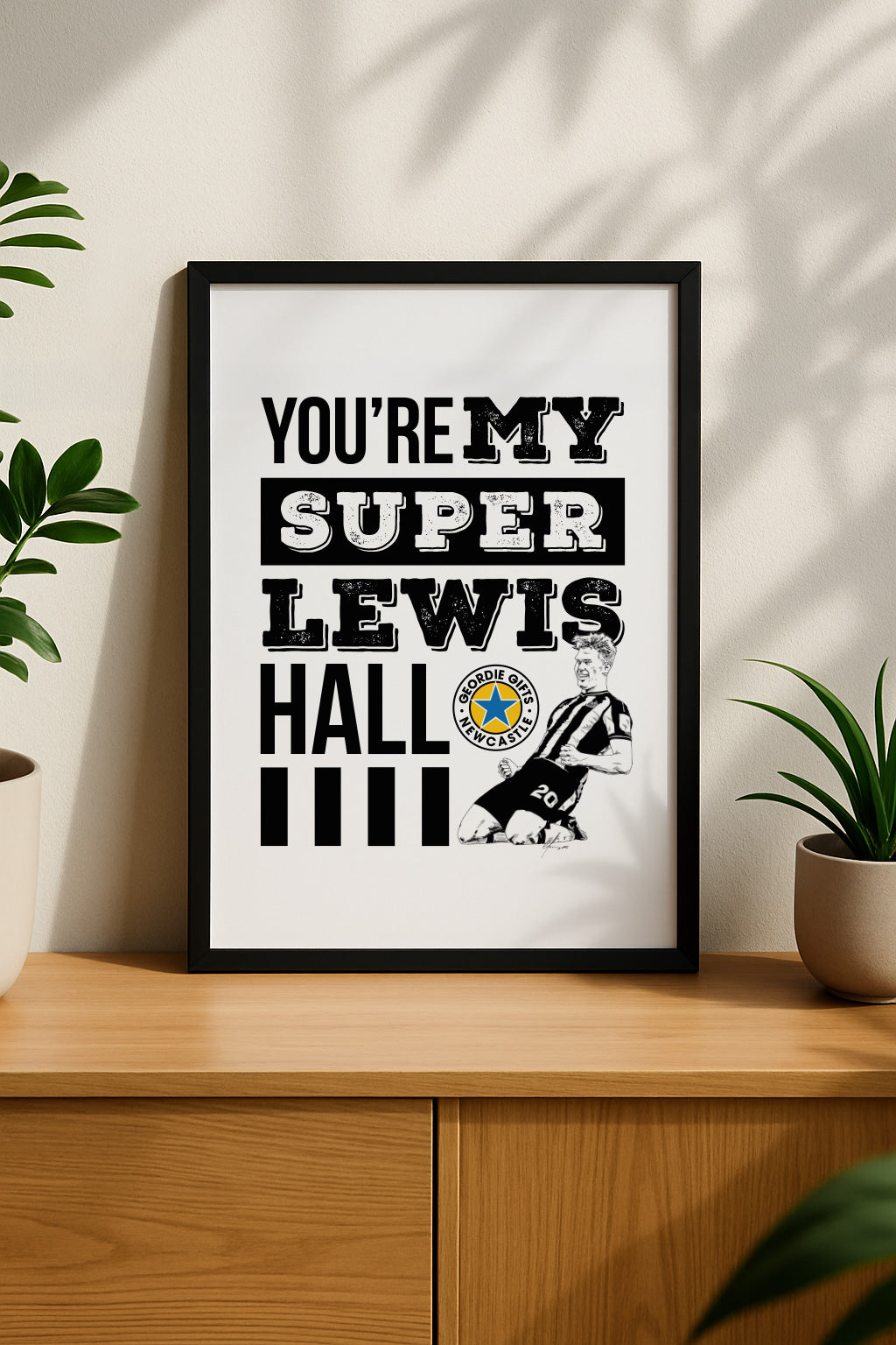 Black framed  on a sideboard"You're My Super Lewis Hall" Newcastle United chant print featuring Lewis Hall goal celebration illustration by Geordie Gifts next to a rose gold alarm clock.   