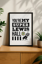 Black framed  on a sideboard"You're My Super Lewis Hall" Newcastle United chant print featuring Lewis Hall goal celebration illustration by Geordie Gifts next to a rose gold alarm clock.   