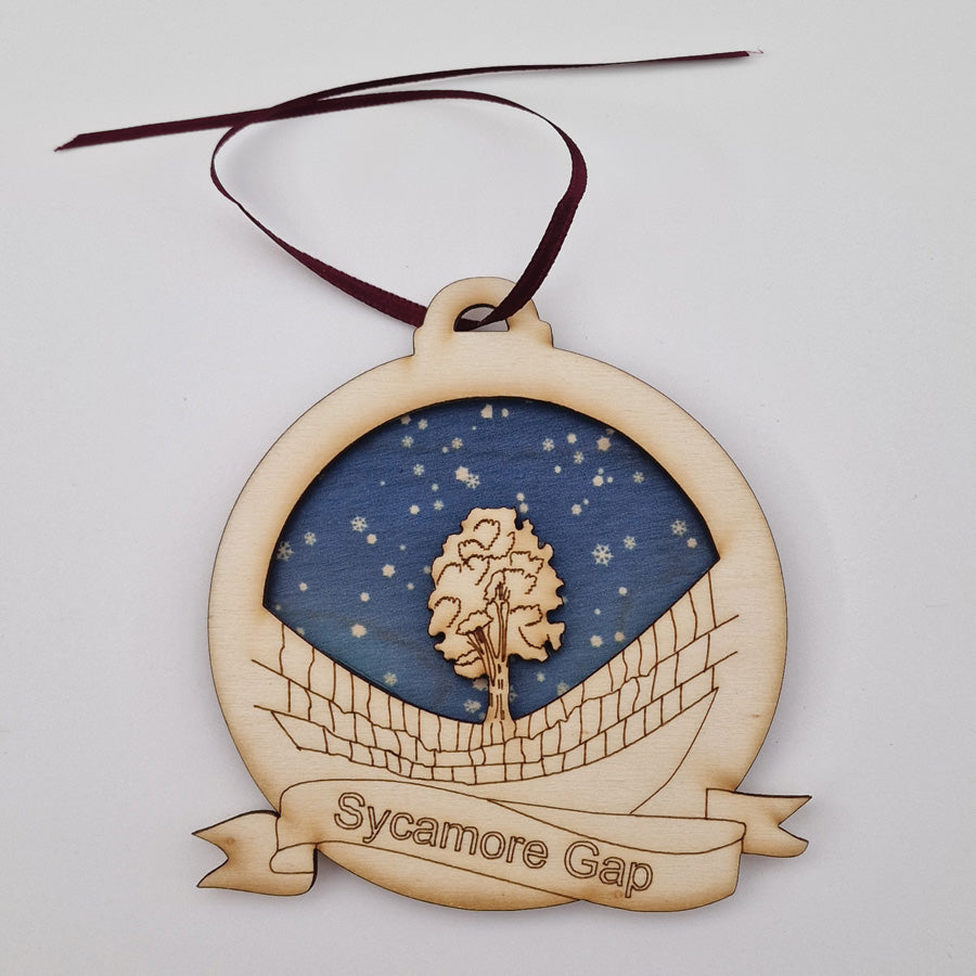 Etched illustration of Sycamore Gap, Northumberland, with a blue snowflake sky background. Geordie Gifts christmas tree wooden bauble decoration